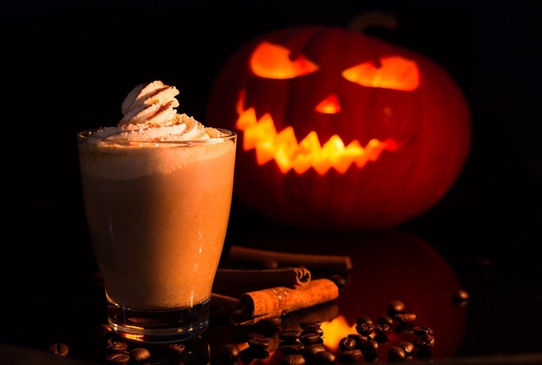 A glass of hot chocolate with a scary carved, cande-lit pumpkin behind it