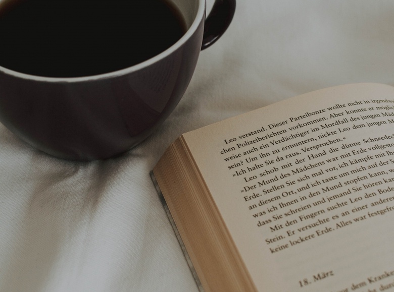 A cup of coffee is shown from bids eye view, positioned next to an open book in warm, calming colours