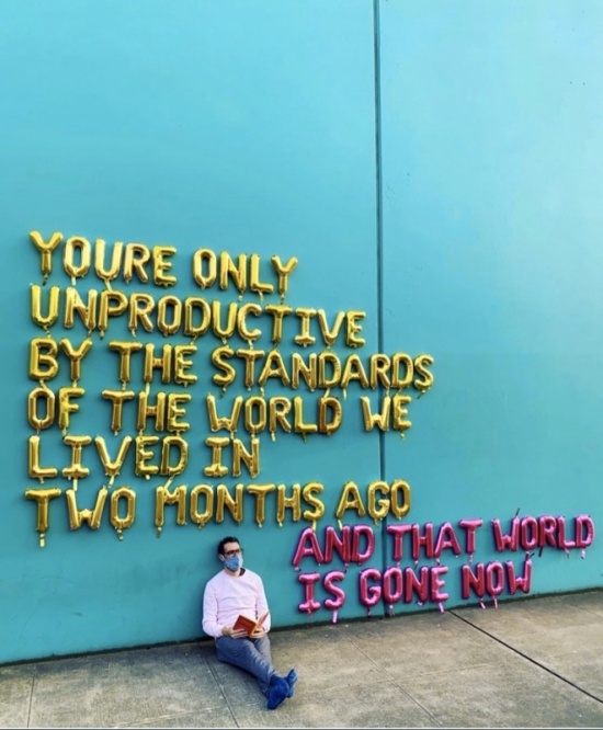 A man leans against a bright blue wall with balloon letters spelling out the words: You're only unproductive by the standards of the world we lived in two months ago. And that world is gone now.