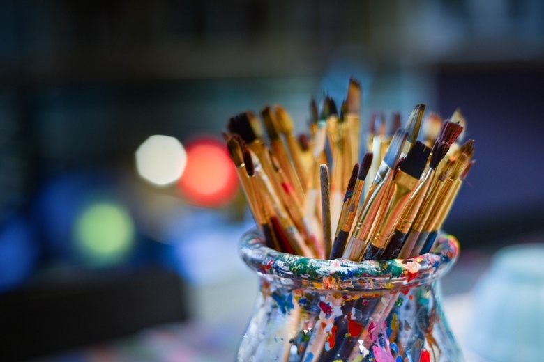 The image shows a glass jar with paintbrushes in it, the jar is spattered with multi-coloured paint and the brushes are all different thicknesses.