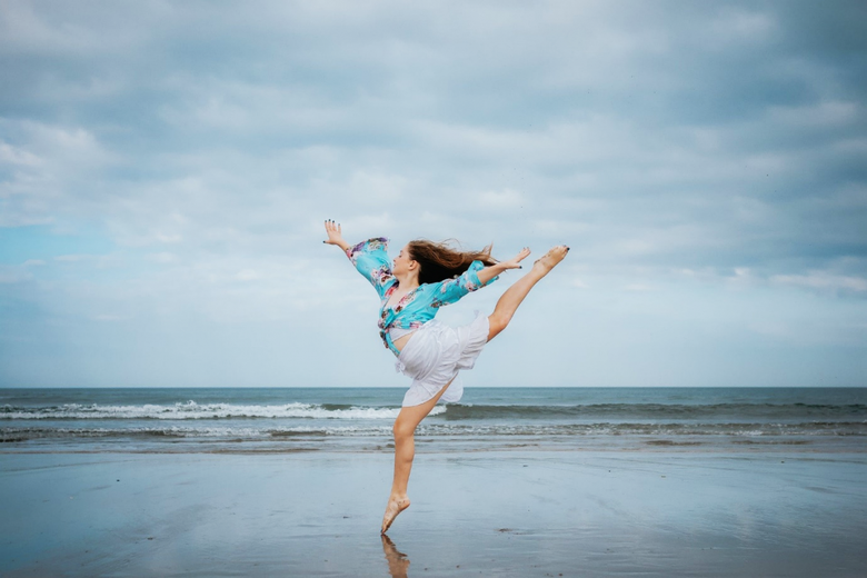 A woman dances on a beach wearing a blue dress. She is jumping into the air with her arms and legs outstretched in a ballet arabesque