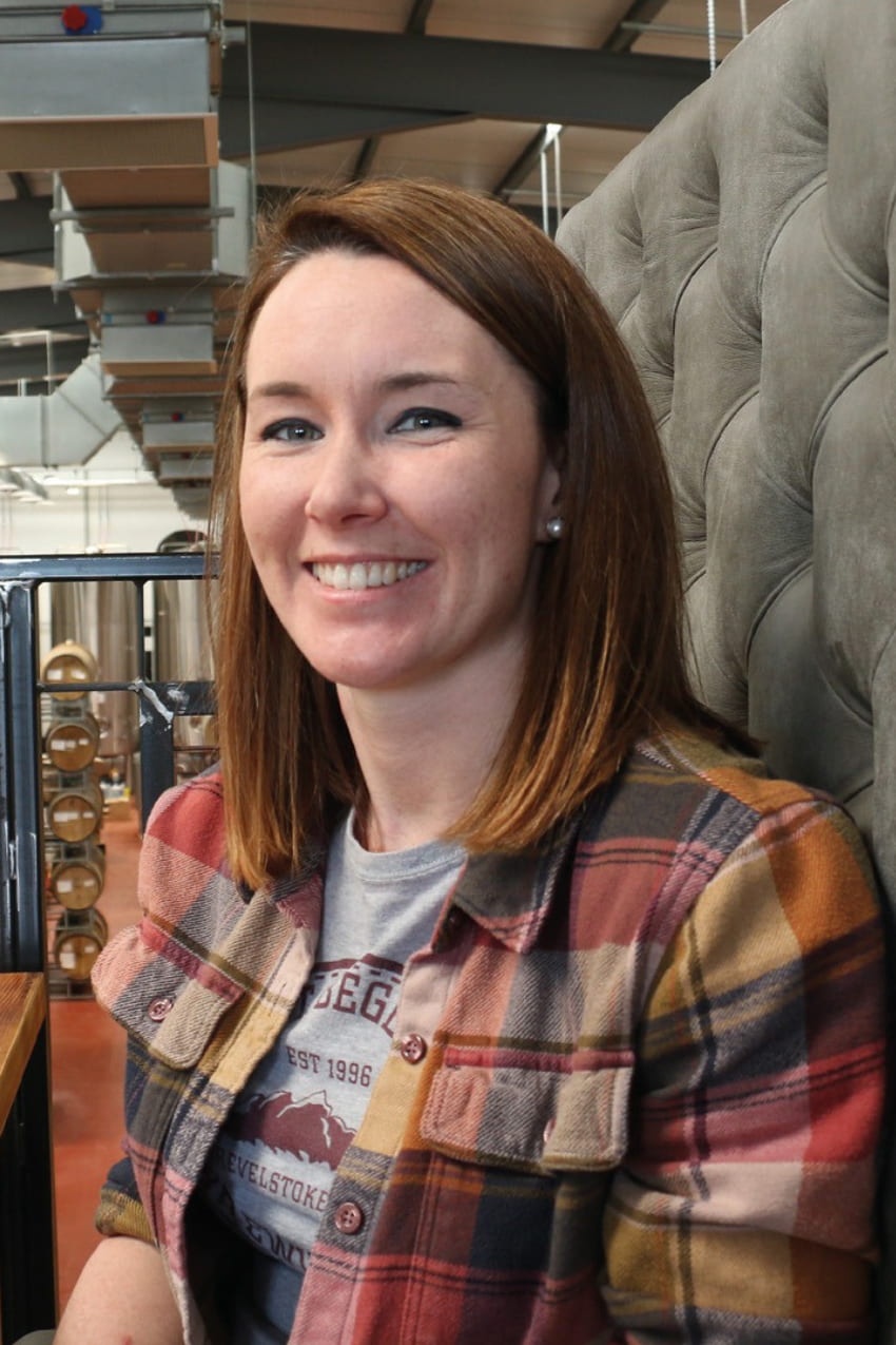 A woman with shoulder-length brown hair and a plaid shirt smiles at the camera