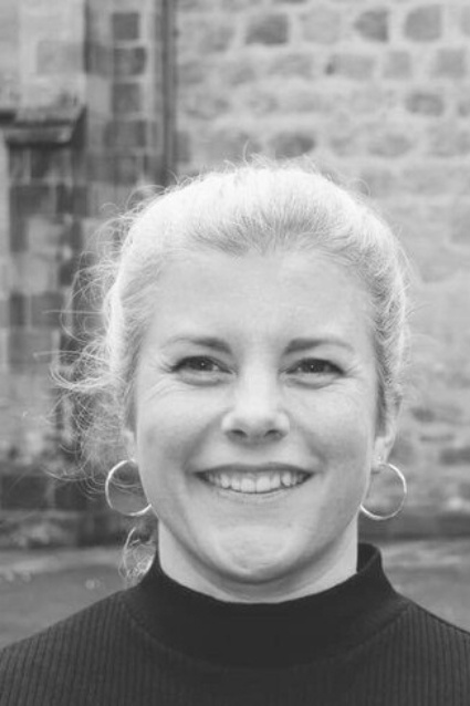 A black and white image of a woman with her hair pulled back, smiling at the camera, with a stone building in the background