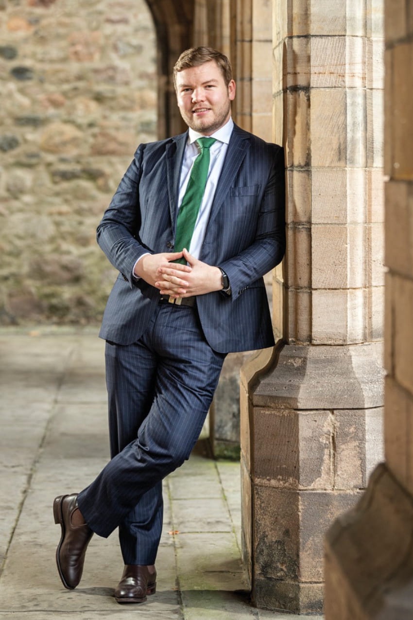 A man in a suit and tie leans against a stone pillar