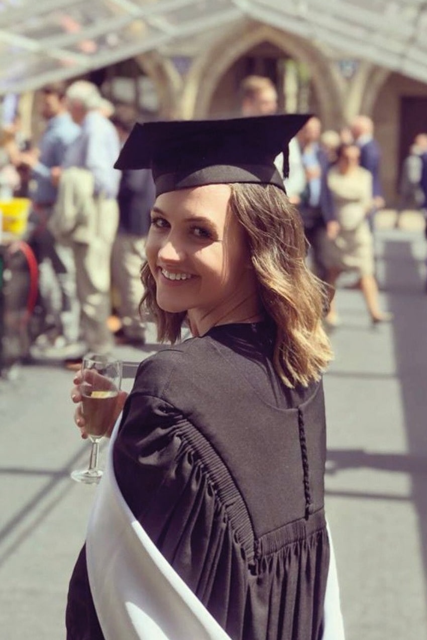 A female university graduate with shoulder-length light brown hair holds a champagne flute and smiles over her shoulder