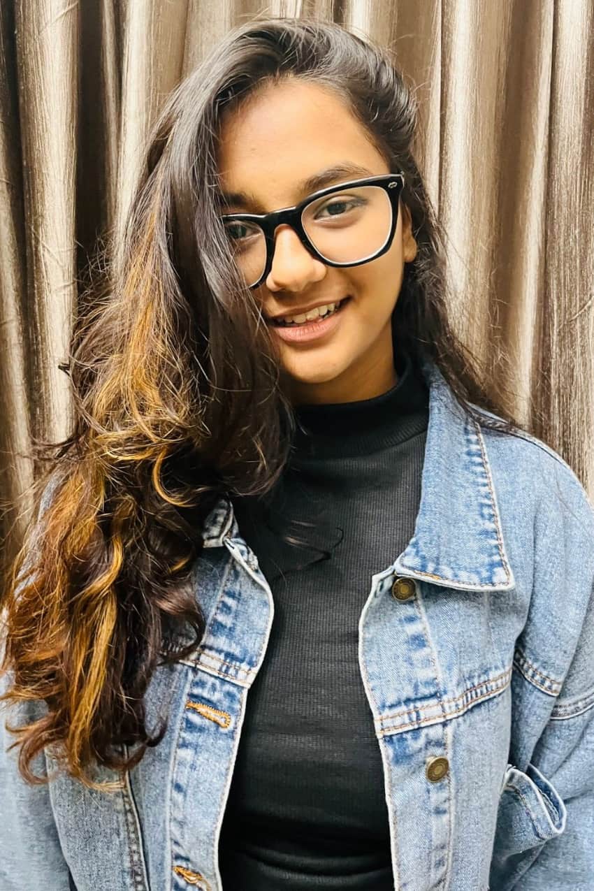 A female student with long dark wavy hair in a jean jacket and glasses smiles at the camera