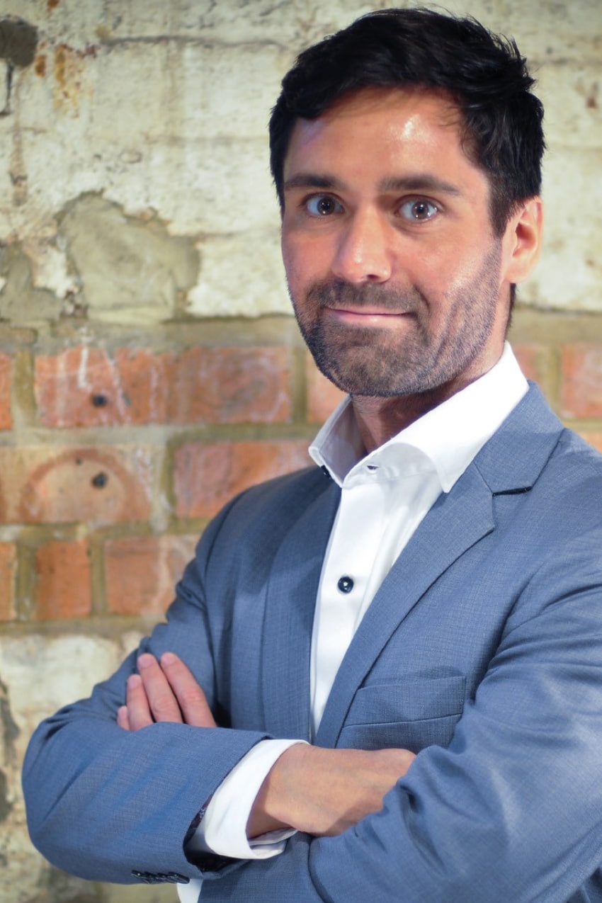 A man with dark hair wearing a light blue-grey suit and white button up shirt, looking towards the camera