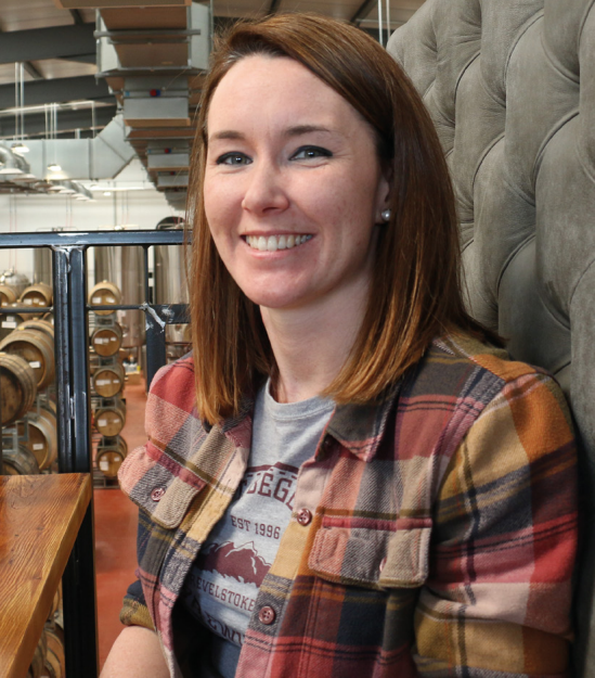 A woman with shoulder-length brown hair and a plaid shirt smiles at the camera