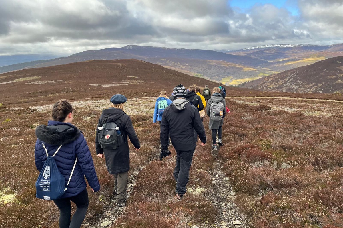 A photo of Aberdeen Philosophers hiking in the Cairngorms. The photo is taken from behind, as students and staff walk down a path toward a mountain range in the distance.