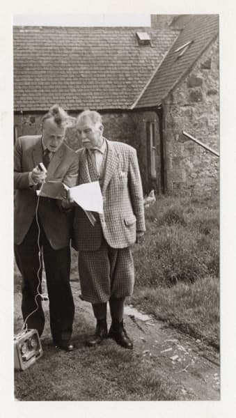 Young man in suit interviewing older man in tweed suit outside.