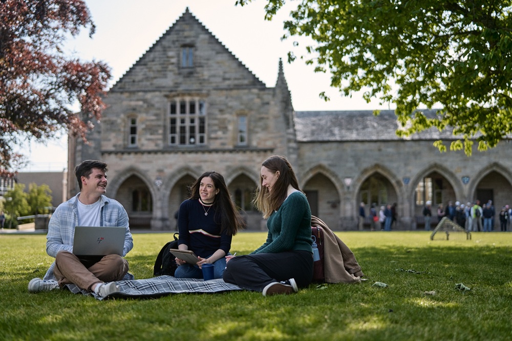 Three students sit on a blanket on Elphinstone Lawn, enjoying a sunny day. Two are using laptops and all are smiling and laughing.