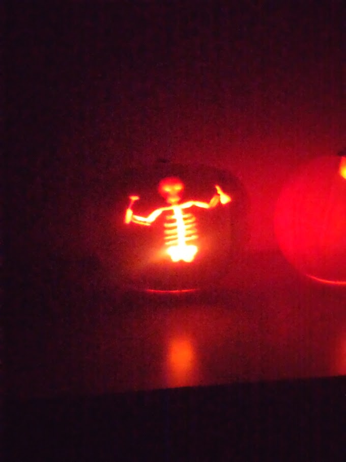 A pumpkin with a carving of a skeleton holding a beaker in one hand and drill in the other. Pumpkin is lit from within in a dark room