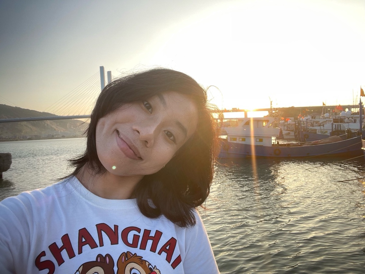 A smiling student in a white cartoon t-shirt, standing in front of water, boats, a bridge, distant hills, and the sunset.