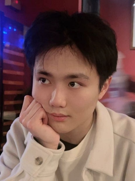 A student sitting indoors, in a room with blue light. Their gaze is up and to the right, and their closed hand rests on their cheek.