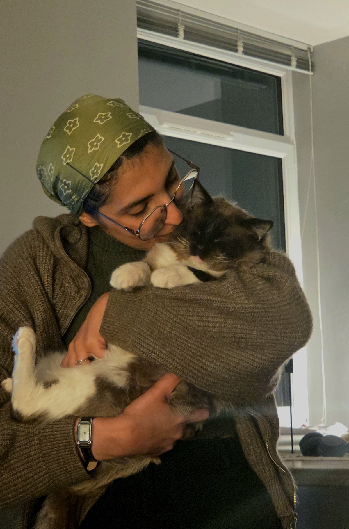 Loujain Alzahrani, in brown clothes, holding a cat in front of a window