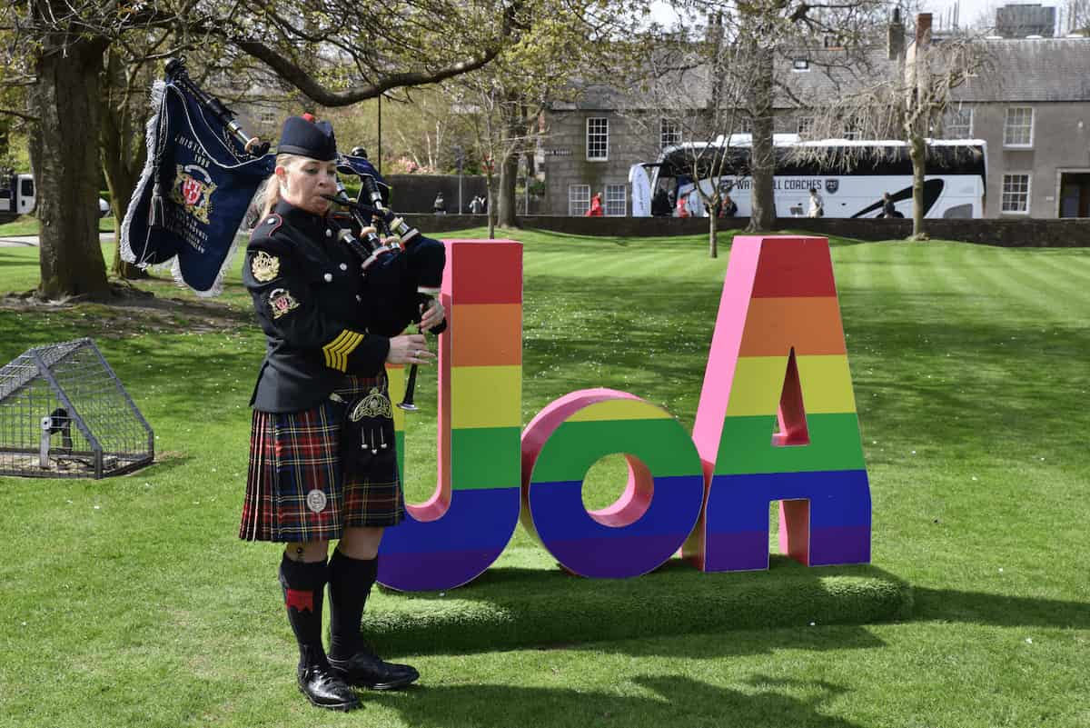 A bagpiper standing next to UOA rainbow letters on campus