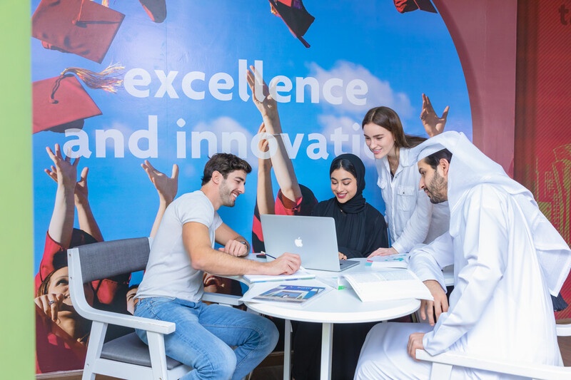 A group of students sitting at a table looking at a laptop, in front of a wall reading