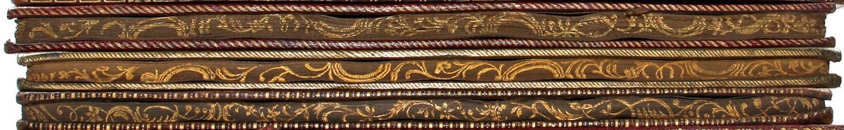Three books piled on top of each other with the edging of the pages visible. The edges are decorated with fine gold details in spirals and leaf figures