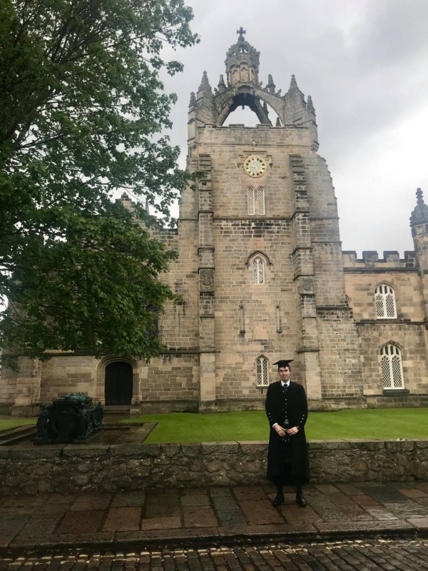 Image shows a young man in kilt and formal graduation robes stands in front of Kings College