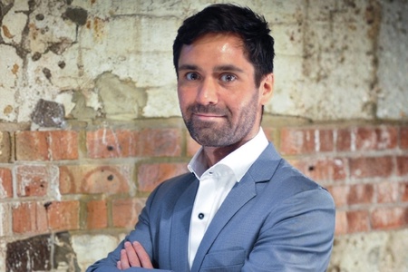A man with dark hair wearing a light blue-grey suit and white button up shirt, looking towards the camera