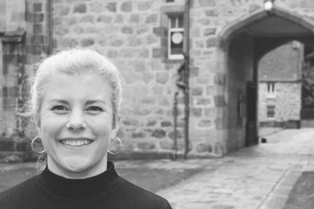A black and white image of a woman with her hair pulled back, smiling at the camera, with a stone building and archway in the background