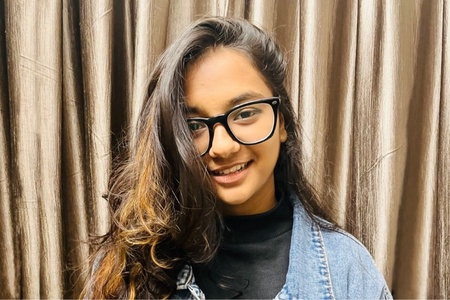 A female student with long dark wavy hair in a jean jacket and glasses smiles at the camera