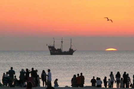 a tall ship on the ocean at sunset watched by silhouetted people on shore