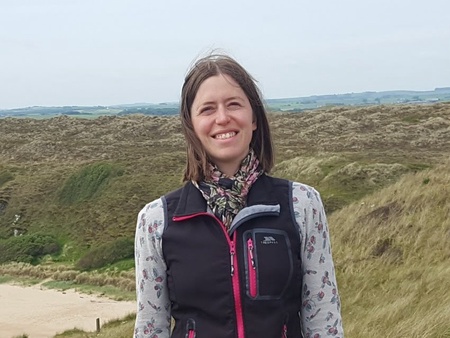 A photo of a lady's head and shoulders. At the beach, wearing a gilet and scarf.