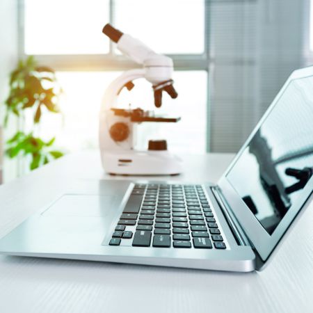 A laptop on a desk with a microscope in the background