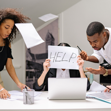 three people at a desk looking busy and demanding input from a person at a laptop who is holding up  sign that says help in front of their face looking overwhelmed