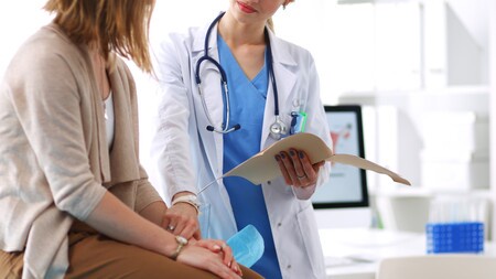 A GP in scrubs and coat converses with a patient