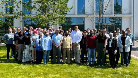 Group photo of postgrad student presenters and attendees and academic staff. Taken outside in front of a building and trees
