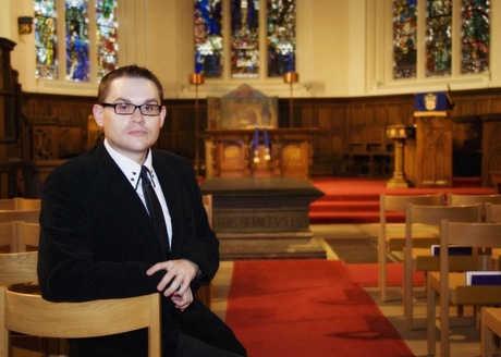 Professor Mealor in a black suit and tie in front of stained glass windows in a chapel