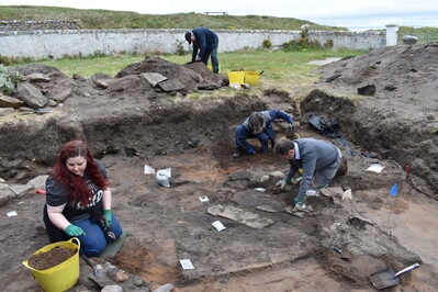 An archaeological excavation site. There is a large trench in the ground an a number of workers are digging for artefacts.