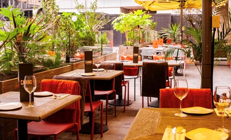 A bright and inviting outdoor restaurant terrace features wooden tables set with wine glasses and black &ldquo;CAMINO&rdquo; menus, surrounded by lush greenery and warm lighting.
