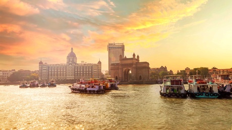 Sunset photo of city scape with The Gateway of India along with Taj Mahal Hotel and boats as seen from the Mumbai Harbour in Mumbai, Maharashtra India with a beautiful warm sky.