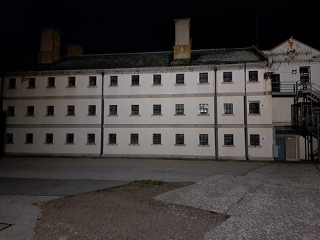 An exterior picture of Peterhead Prison Museum at night. The museum is surrounded by darkness, with a light illuminating the building. There are many small windows covered with bars.