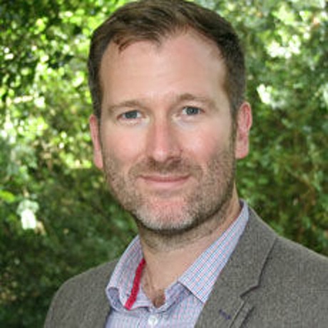 Photo of man smiling at camera. Greenery in background.