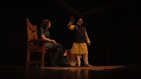 A man gestures while a woman sitting on a wooden chair looks on
