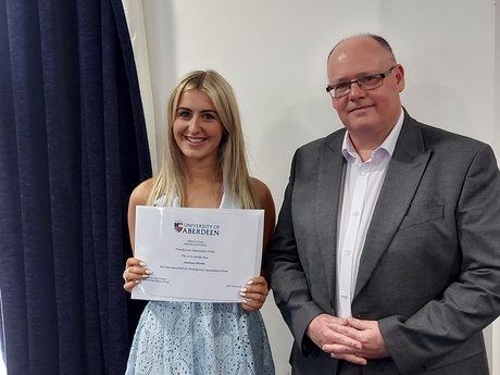 Photograph of Prize winner Joanna Sheils, holding their certificate, alongside Head of Law School Prof Greg Gordon