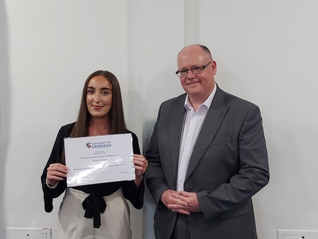 Photograph of Prize winner Katie Clark, holding their certificate, alongside Head of Law School Prof Greg Gordon