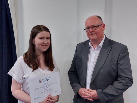 Photograph of Prize winner Sarah McNeill, holding their certificate, alongside Head of Law School Prof Greg Gordon