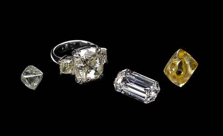 Against a black background sits two large un cut diamonds, one emerald cut diamond and a ring with a large square diamond and two smaller diamonds on the side
