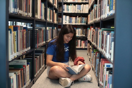 Image of a student sitting on the floor reading, surrounded by bookshelves. Photo by Ludovic Delot on Unsplash