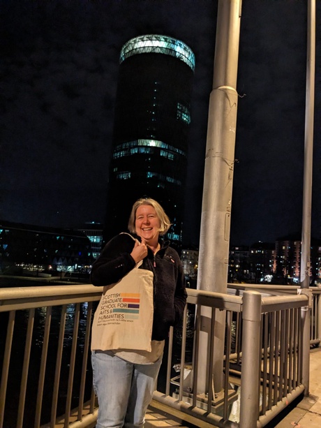 Sian MacGregor, standing in front of a river in a city in the evening, smiling, with a tall illuminated building visible in the background.