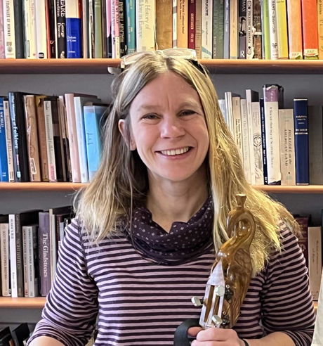 Dr Frances Wilkins, smiling and holding a musical instrument in front of bookcases, which are filled with books.