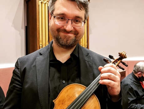 Dr Aaron McGregor in a black shirt and suit, holding a stringed instrument, in front of a pipe organ.