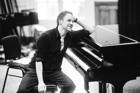 Gareth Williams, in a blakc and white image, leaning against a piano in a quiet room