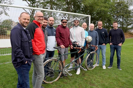 Ten men standing beside a tandem bike in front of a football goal