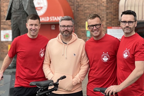 Four men standing in front of a tandem bike outside Pittodrie stadium
